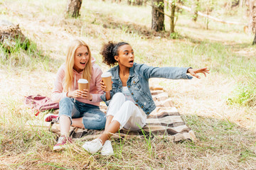 two amazed multiethnic friends sitting on plaid with paper cups of coffee and looking away