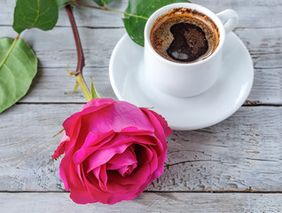 Romantic background: a cup of coffee standing on a wooden textured background with a pink rose lying next to it.