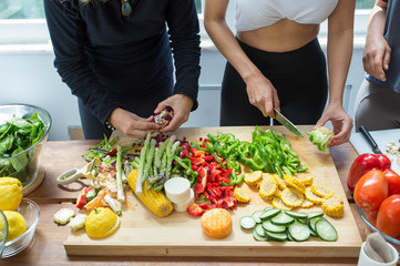 Closeup of women peeling and cutting vegetables and fruits. Ladies cooking salad at table with fresh sweet pepper, asparagus and pomegranate. Healthy cooking and organic food concept. Front view.