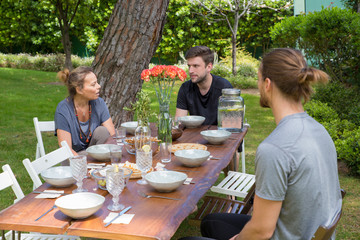 Serious people having lunch at table in backyard. Woman and men chatting, eating and sitting at wooden table with plates, food and flowers and green plants in background. Summer and meal concept.