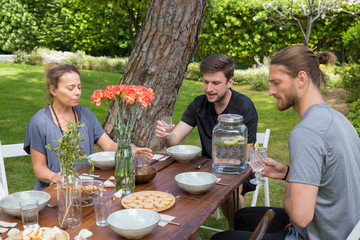 Serious people having lunch at wooden table in patio. Woman and men chatting, eating and sitting at table with plates, food, flowers and green plants in background. Summer and breakfast concept.