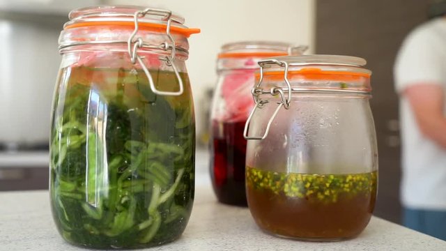 handheld shot of fermented wild garlic, fermented red berries, pickled wild garlic flowers and raspberry vinegar in jars in a kitchen with the chef working in the kitchen in the blurred background.