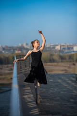 Fototapeta na wymiar Ballerina in a tutu posing near the fence. Beautiful young woman in black dress and pointe dancing outside. Gorgeous ballerina demonstrates amazing stretching.
