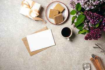 Fototapeta na wymiar Minimal home office desk workspace with white paper card, kraft envelope, coffee cup, cookies, lilac flowers bouquet, perfume bottle, glasses. Flat lay, top view social media background.