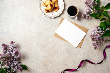 Flatlay of kraft envelope and blank paper card with copy space mock up. Top view home office desk workspace decorated with lilac flowers, coffee cup, cookie, purple ribbon. Women desk concept.