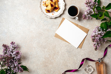 Flatlay of kraft envelope and blank paper card with copy space mock up. Top view home office desk workspace decorated with lilac flowers, coffee cup, cookie, purple ribbon and perfume bottle