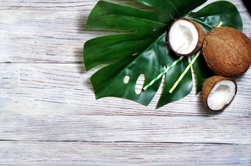 coconuts and tropical leaf of monstera plant with a paper straw cocktail on white wooden background. Flat layer, top view, copy space. Healthy cooking. Creative concept of healthy eating. horizontal