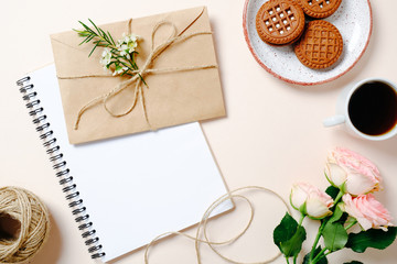 Flat lay home office desk. Female workspace with kraft paper envelope, blank paper notebook, coffee cup, cookies and roses flowers on pink background. Top view feminine background
