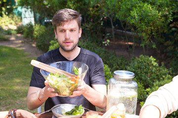 Portrait of serious young man passing salad at lunch outdoors. Guy having meal with family outside. Brunch concept