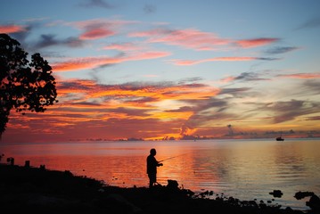 Silhouette of an unrecognizable man casting a fishing pole in the waters with a blood-red sunset in the background.