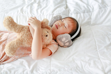 Cute little Asian girl wearing winter ear muffs and embracing teddy bear lying on the bed at home.