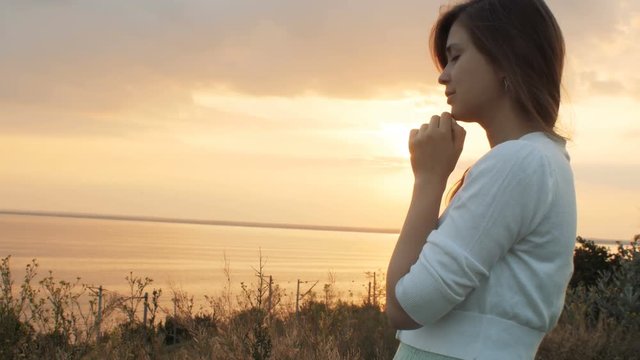 face profile of young beautiful woman turning to God in nature at sunset, the girl praying folded her hands at the chin, concept of religion