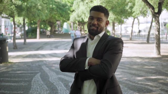 Confident bearded businessman standing with crossed arms. Handsome African American man wearing formal suit posing in summer park. Concept of confidence