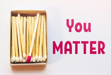 White matches and one match of crimson color in a box. Difference, self respect and acceptance concept. Motivational phrase on the image. Opposition, uniqueness, individualism concept. Not alike.