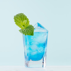 Exotic alcohol cold blue cocktail with curacao liquor, ice and green mint in rich wet shot glass on soft light pastel green background.