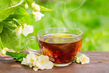 Glass cup with hat tea and jasmine twig on wooden table, on blurred background