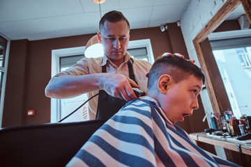 Small school boy is getting trendy haircut from expirienced barber at modern barbershop.