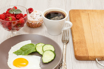 Plate with fried egg, cut cucumber and leaf of basil, glass bowl with raspberries, cup of coffee, fork and muffin.