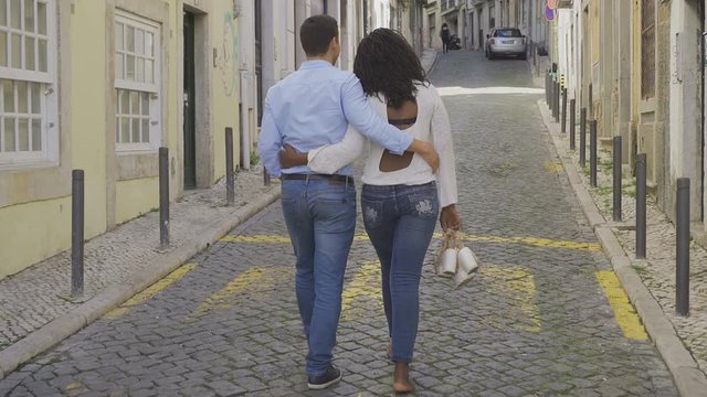 Back view of young couple strolling on paved street. Smiling Caucasian man and barefoot African American woman walking on street. Relationship concept