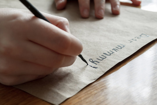 A small girl writes a letter to dear mummy by black paint on brown paper. Closeup, selective focus