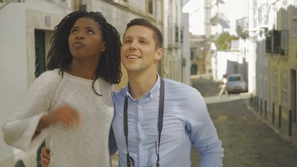 Happy couple of tourists looking and pointing up. Smiling African American woman and Caucasian man with digital photo camera hugging outdoor. Travel concept