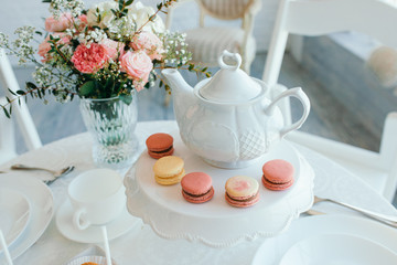 Fototapeta na wymiar Creative spring composition. Elegant sweet dessert macarons, cup of tea or coffee and beautiful pastel colored beige and living coral flowers bouquet on white marble background