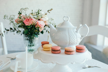 Creative spring composition. Elegant sweet dessert macarons, cup of tea or coffee and beautiful pastel colored beige and living coral flowers bouquet on white marble background