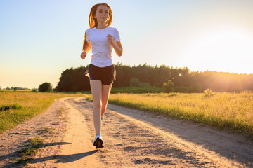 Young fitness girl in a white t-shirt running on the dirt road across the field in the sunrise.