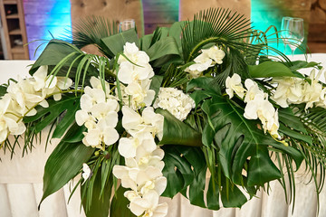 Lush floral arrangement of orchids and monstera leaves on wedding table. Wedding presidium in...