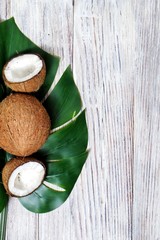 two halves of coconut and one whole coconut with a leaf of tropical monstera plant on a white wooden background. Flat layer, top view, copy space. Creative concept of healthy eating. vertical