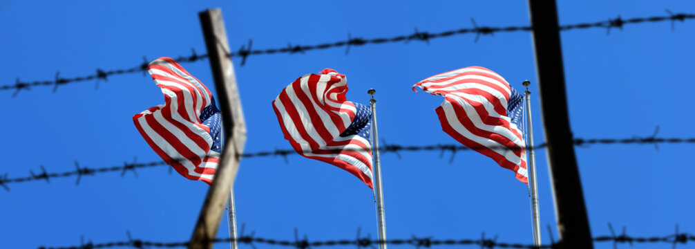Conceptual image of waving American flags at tall pole and barbed wire fence over blue sunny sky