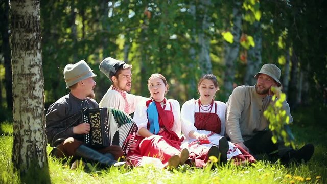 People in traditional Russian clothes sit on the lawn and singing by the accordion