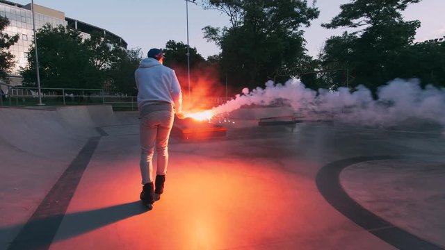 Aggressive Inline roller skater doing tricks in concrete skatepark with red burning signal flare outdoors, slow motion