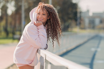 Young happy attractive woman posing on a fence near a running track