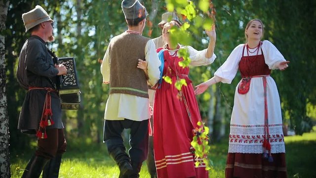 People in traditional Russian clothes are dancing on the field holding hands - one of them plays the accordion music