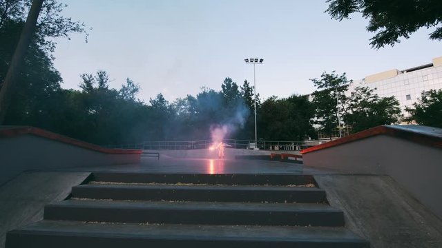 Aggressive Inline roller skater doing tricks in concrete skatepark with red burning signal flare outdoors, slow motion
