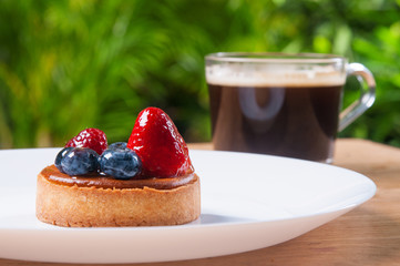 Closeup of beautiful mini tart with berries and cup of coffee. Delicious pastry with strawberries and blueberries on plate on table with blurred green view in background. Breakfast concept.