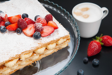 Close-up of appetizing Napoleon cake with berry and sweet powder. Cappuccino in cup and cake stand with decorated cake on table. Breakfast in coffee shop concept