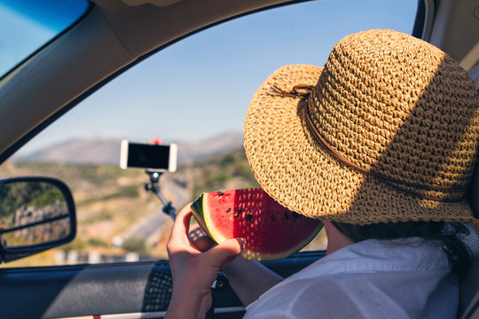 Blogger girl with slice of watermelon and  straw hat taking selfie picture or video using smartphone and selfie stick inside of car.