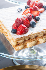 Close-up of Napoleon cake with custard cream and ripe berries as topping. Tasty dessert on transparent cake stand. Bakery concept