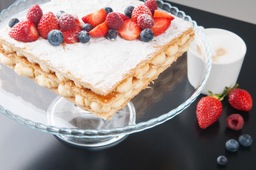 Close-up of sweet cake on glassy stand in cafeteria. Coffee cup and fresh berries in background. Dessert concept