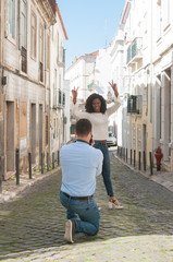 Fototapeta na wymiar Man taking photo of playful black woman outdoors. Woman showing victory signs and man standing back to camera in city street. Tourism concept.