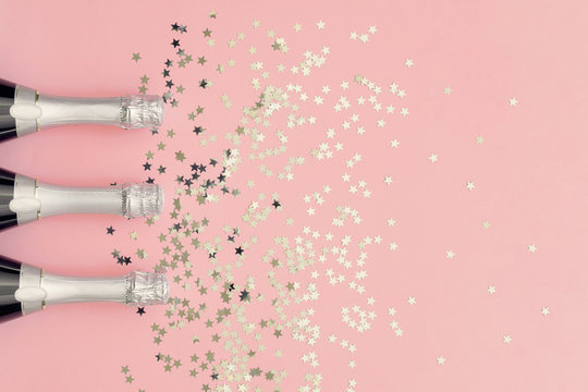 Three Champagne bottles with silver confetti stars on bright coral background. Copy space, top view.