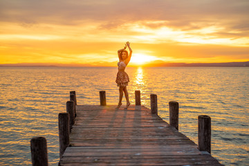 Happy woman enjoying the summery sunset at the lake on her vacation. The woman in bra and a short summer skirt laughs and puts her arms in the direction of the sky