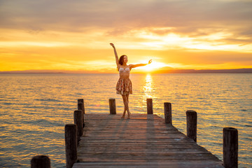 Fototapeta premium Happy woman enjoying the summery sunset at the lake on her vacation. The woman in bra and a short summer skirt laughs and puts her arms in the direction of the sky