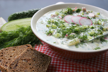 Okroshka is a traditional Russian summer cold soup in a ceramic bowl. Ingredients potatoes, radishes, cucumbers, dill, eggs, kefir (yogurt), sausage. Summer yogurt cold soup on a wooden table. Selecti
