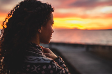 Thoughtful evening mood with a young Afro American woman standing on the promenade by the lake and looking towards the water and the setting sun. Burning sky