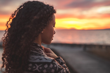 Thoughtful evening mood with a young African American woman standing on the promenade by the lake and looking towards the water and the setting sun. Burning sky
