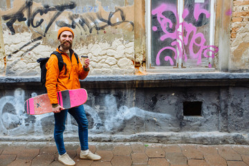 Hipster positive beard man traveler with coffee cup holding longboard in his hand in orange hat and...