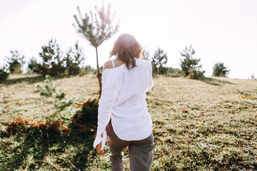 Beautiful young woman in white collecting grass at the rural sunny landscape background in summer. Tender happy woman in wild field enjoying nature. Back view